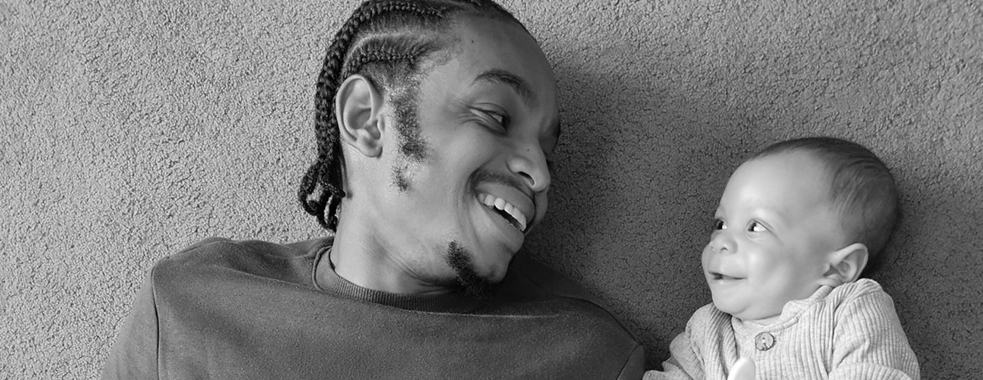 Black and white photo of a Black man and a baby lying on their backs, laughing with each other